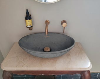 May include: A gray oval bathroom sink with a brass faucet and drain, set on a marble-topped vanity. A matching brass soap dispenser is mounted on the wall. The sink has the name "Voglio" written on it.