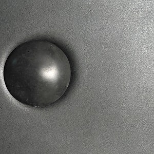 Könnte beinhalten: Nahaufnahme eines schwarzen, runden Metallknopfes auf einer silbernen, strukturierten Oberfläche.