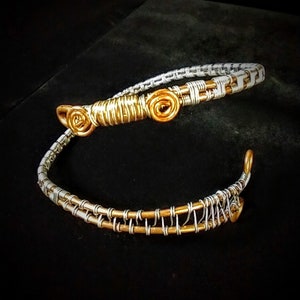 Puede incluir: Dos pulseras de alambre, una plateada y otra dorada, están entrelazadas. La pulsera dorada tiene tres espirales decorativos.