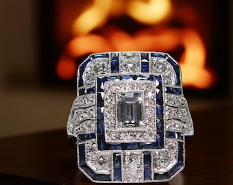 May include: A rectangular, Art Deco-style ring with a large, emerald-cut center stone. The ring features a geometric design with blue sapphire accents and smaller diamonds, set in a silver or white gold band. The ring is displayed against a blurred background.