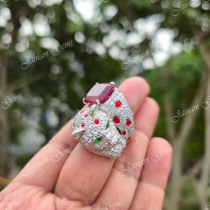 May include: A silver ring with a panther head design, set with a large, rectangular red gemstone. The ring is embellished with small, clear stones, and red and green accents, offering a detailed and luxurious look.