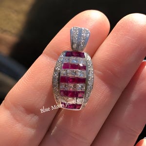 May include: A silver pendant with a red ruby and diamond design. The pendant is oval shaped and has a bail at the top. The ruby stones are set in a row down the center of the pendant, with diamonds set on either side.