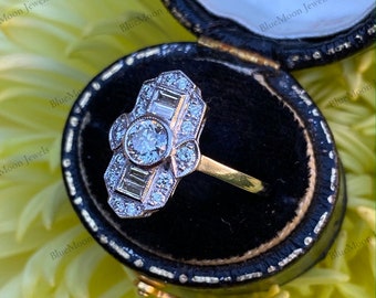 May include: A vintage-style diamond engagement ring with a large round center stone and smaller baguette and round diamonds set in a gold band. The ring is in a black velvet ring box.