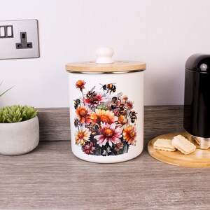 May include: White canister with a wooden lid and knob. The canister has a floral and bee design with orange, pink, and yellow flowers. The canister is on a wooden surface.