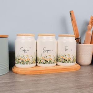 Può includere: Tre barattoli da cucina in ceramica bianca con coperchi in legno, etichettati "Coffee", "Sugar" e "Tea". Ogni barattolo presenta un motivo floreale con fiori gialli e verdi. I barattoli sono su un vassoio di legno.