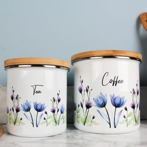 May include: Two white enamel canisters with bamboo lids, labelled "Tea" and "Coffee." Each canister features a floral design with blue and purple flowers and green leaves. The canisters are cylindrical and have a silver rim.