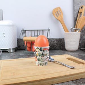 May include: A ceramic egg cup featuring a colourful illustration of houses and trees. An orange egg sits inside the cup. A wooden chopping board and a silver spoon are also in the image.