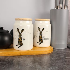 Op de afbeelding: Twee witte keramische potten met houten deksels, versierd met een hazenillustratie. Eén pot heeft de tekst "TEA", de andere "HOT CHOCOLATE". Ze staan op een houten dienblad, met een zwarte theepot en een grijze keukengerei houder op de achtergrond.