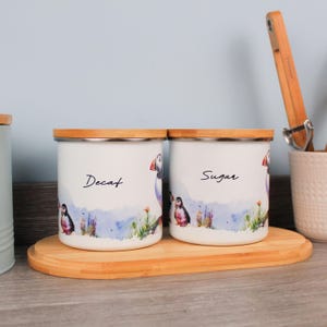 May include: Two white storage jars with bamboo lids, labelled "Decaf" and "Sugar." Each has a watercolour design of puffins, grass, and a blue and purple landscape. The jars sit on a wooden tray.