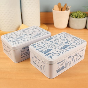 Può includere: Due scatole rettangolari in metallo bianco con illustrazioni di utensili blu sui coperchi e sui lati. Le scatole sono su una superficie di legno.