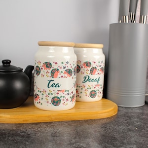 Op de afbeelding: Twee crèmekleurige keramische potten met houten deksels, versierd met een bloemen- en egelsmotief. Eén pot is gemarkeerd met "Tea" en de andere met "Decaf". Een zwarte theepot en een grijze keukengereihouder zijn ook zichtbaar.