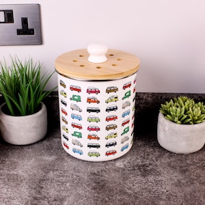 May include: A white ceramic canister with a bamboo lid and a white knob. The canister is decorated with colourful illustrations of vintage camper vans. The lid has several small holes. The canister is on a countertop.