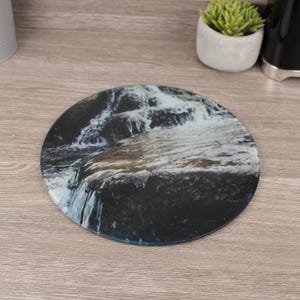 May include: Circular glass coaster depicting a waterfall scene in blue, white, and brown hues. This coaster is designed to protect surfaces from drink condensation. The coaster has a diameter of approximately 10 cm.