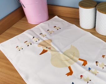 Könnte beinhalten: Weißes Küchentuch mit einer großen Cartoon-Gans mit Kochmütze, umgeben von kleineren Gänsen mit Hüten und Schals. Ein rosa Eimer und zwei weiße Behälter mit Holzdeckeln befinden sich im Hintergrund.