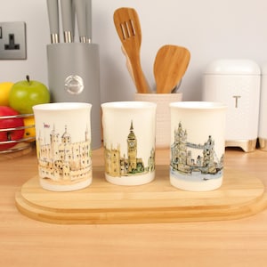 Peut inclure: Trois mugs en céramique blanche décorés de monuments londoniens : la Tour de Londres, Big Ben et le Tower Bridge. Les illustrations sont dans des tons doux, posés sur une planche en bois. Articles de cuisine.