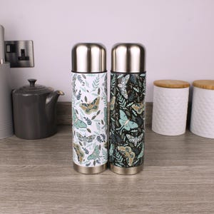 Peut inclure: Deux bouteilles isothermes cylindriques en acier inoxydable avec des bouchons en argent métallique. L'une présente un fond blanc avec un motif répété de papillons de nuit et de feuilles vertes, l'autre un fond noir avec le même motif.