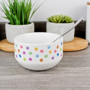 May include: White ceramic bowl with a pattern of colourful polka dots in pink, purple, blue, green, and orange. A stainless steel spoon is inside the bowl. The bowl is on a wooden surface.