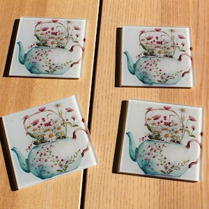 May include: Set of four square glass coasters featuring a teapot filled with white and pink flowers. The teapot is painted in a light blue and white colour scheme with a floral pattern.