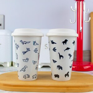 May include: Two white ceramic travel mugs with lids. One mug features a black and white geometric pattern of origami animals. The other mug features a black and white geometric pattern of origami birds.