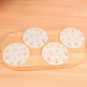 May include: Four circular glass coasters with a white base, decorated with orange, yellow, and pink starburst patterns. They are displayed on a wooden serving tray.