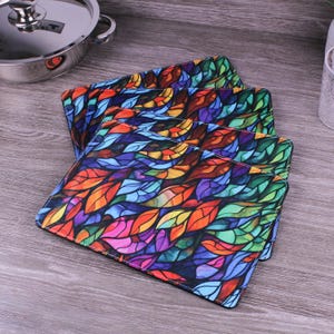 May include: A set of rectangular placemats featuring a vibrant, multicoloured stained-glass style pattern of overlapping leaves in shades of red, orange, yellow, blue, purple, and green. The placemats have stitched edges and a smooth, flat surface.