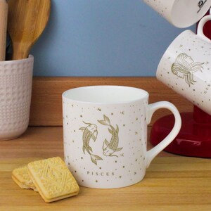 May include: A white ceramic mug with a gold Pisces design, featuring two fish and the word "PISCES". The mug has a curved handle. Other mugs and biscuits are also visible.