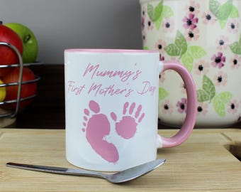 Puede incluir: Taza de cerámica blanca con borde y asa de color rosa. La taza tiene un diseño rosa de dos huellas de pies de bebé y el texto "Mummy's First Mother's Day."