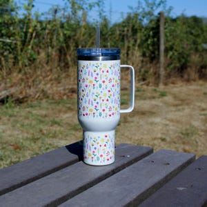 May include: A white stainless steel tumbler with a handle and a clear straw, featuring a repeating pattern of small, colourful wildflowers in shades of yellow, pink, red, and blue. The cup has a blue lid and a tapered base design.