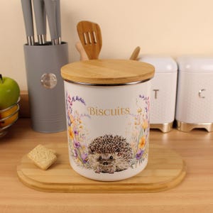 May include: White biscuit jar with a wooden lid, decorated with a hedgehog and floral pattern. The word "Biscuits" is written on the front. Other kitchen items, such as a knife block and wooden utensils, are visible.