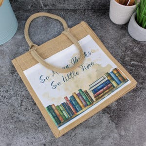 Puede incluir: Una bolsa de lona marrón con un panel blanco que presenta una ilustración de acuarela de una estantería llena de libros coloridos. El texto "So many books, so little time" está impreso en el panel.