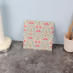 Può includere: Piastrella decorativa quadrata con motivo floreale. Il design presenta fiori rosa, foglie verdi e steli marroni su sfondo chiaro. La piastrella è posizionata su una superficie grigia con una parete azzurra sullo sfondo.