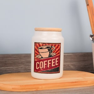 Op de afbeelding: Een witte keramische koffiepot met een houten deksel. De voorkant heeft een retro-afbeelding van een koffiekopje en het woord "COFFEE" in een rood en zwart spandoek. De pot staat op een houten snijplank.
