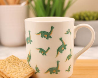 Op de afbeelding: Een crèmekleurige keramische mok met een herhalend patroon van groene en gele langnekdinosaurussen. De mok heeft een gladde afwerking, een gebogen handvat aan de rechterkant en een cilindrisch lichaam. Een vierkant, getextureerd koekje ligt ernaast.