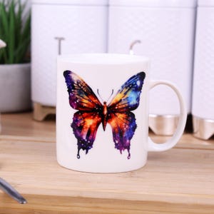 Peut inclure: Tasse en céramique blanche ornée d'un motif de papillon coloré. Le papillon présente un motif galactique avec des nuances de violet, d'orange et de bleu. La tasse a une anse incurvée et est posée sur une surface en bois.