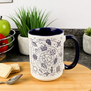 Puede incluir: Taza de cerámica blanca con interior y asa azul marino, decorada con un diseño en línea azul de elementos espaciales. La taza muestra planetas, cohetes y astronautas. Está sobre una tabla de madera.