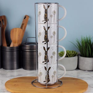 Puede incluir: Un juego de cuatro tazas de cerámica blanca apiladas en un estante de metal cromado. Cada taza presenta una ilustración de liebre marrón con acentos de diente de león. Las tazas se exhiben sobre una superficie de madera.