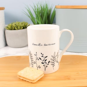 Può includere: Una tazza di ceramica bianca con le parole "Aromatic Americano" e illustrazioni floreali nere. Un piccolo biscotto quadrato è posto davanti alla tazza su un tagliere di legno. La tazza ha un manico curvo.