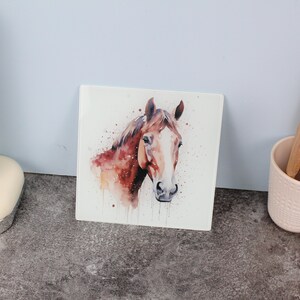 Può includere: Piastrella quadrata in ceramica con un dipinto ad acquerello della testa di un cavallo. Il cavallo ha una criniera e un mantello marroni con segni bianchi sul viso. Lo sfondo è bianco con macchie marroni, su una superficie grigia.