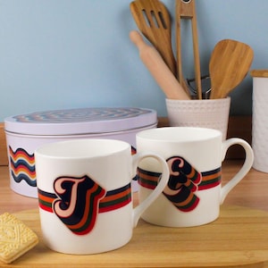 Può includere: Due tazze in ceramica bianca con una grande lettera "J" in un font retrò, accentuata da una fascia a righe arancioni, verdi e blu navy. Una latta e un biscotto abbinati sono sullo sfondo. Articoli per la cucina.