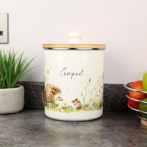May include: A cream-coloured compost bin with a wooden lid and a cream knob. The bin is decorated with a watercolour illustration of a hedgehog and a mouse in a field of wildflowers. The word "Compost" is written in script.