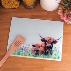 May include: A white glass chopping board featuring a brown Highland cow and calf design. The cows are standing in a field of green grass and wildflowers. The chopping board is on a wooden surface.