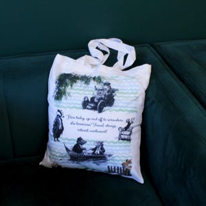 Puede incluir: Una bolsa de tela blanca con una ilustración impresa de animales antropomórficos en varias escenas de viaje, incluyendo un coche y un barco. El texto dice: "Here today, up and off to somewhere else tomorrow! Travel, change, interest, excitement!"