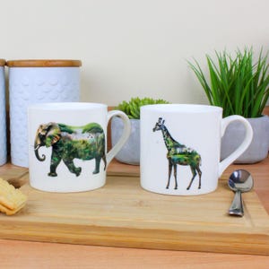 May include: Two white ceramic mugs. One depicts an elephant with a landscape design, the other a giraffe with a similar landscape. Both mugs have a curved handle. They are displayed on a wooden tray.