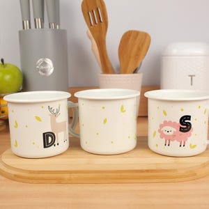 May include: Three cream enamel mugs with animal designs and letters. One mug has a deer with the letter "D", another a sheep with "S", and the third a leaf pattern. The mugs are on a wooden tray, ideal for tea or coffee.