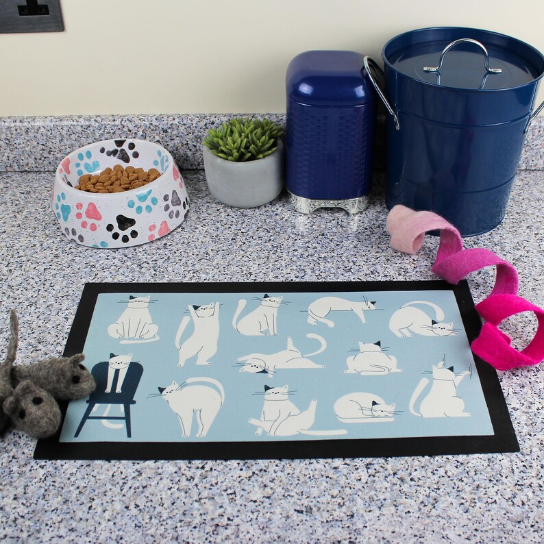 May include: A blue and white cat-themed non-slip kitchen mat with a black border. The mat features a variety of white cats in different poses on a light blue background.