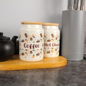 Può includere: Due barattoli in ceramica color crema con coperchi in legno, etichettati "Coffee" e "Hot Choc." I barattoli presentano un motivo floreale con dettagli marroni e arancioni. Sono su un vassoio di legno accanto a una teiera nera e un portautensili grigio.