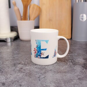Può includere: Tazza in ceramica bianca con la lettera "E" in acquerello blu, decorata con coralli e conchiglie. La tazza ha un manico curvo ed è appoggiata su un piano di lavoro della cucina, con utensili di legno e un tagliere sullo sfondo.