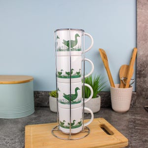 May include: Four white ceramic mugs with green duck designs, stacked in a chrome rack. The mugs are on a wooden board. The background is light blue. The mugs are suitable for tea or coffee.