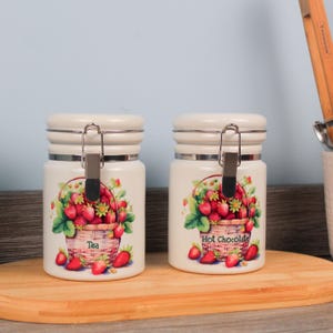 May include: Two cream ceramic storage jars with silver metal clasps. Each jar has a strawberry basket design and the words "Tea" and "Hot Chocolate". The jars are on a wooden chopping board.