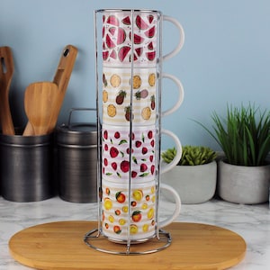 Può includere: Un set di quattro tazze impilate con disegni di frutta, tra cui anguria, ananas, lamponi e arance. Le tazze sono tenute in un supporto in metallo cromato e poggiano su un tagliere di legno. Le tazze hanno manici bianchi.